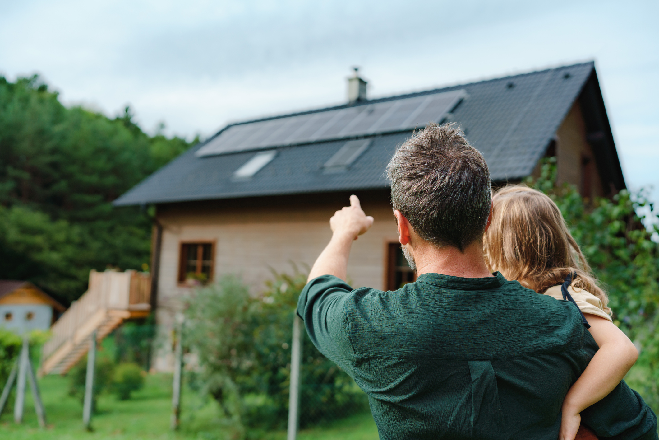 Erwachsener mit Kind blickt auf Wohnhaus mit Photovoltaikanlage und zeigt in Richtung Dach