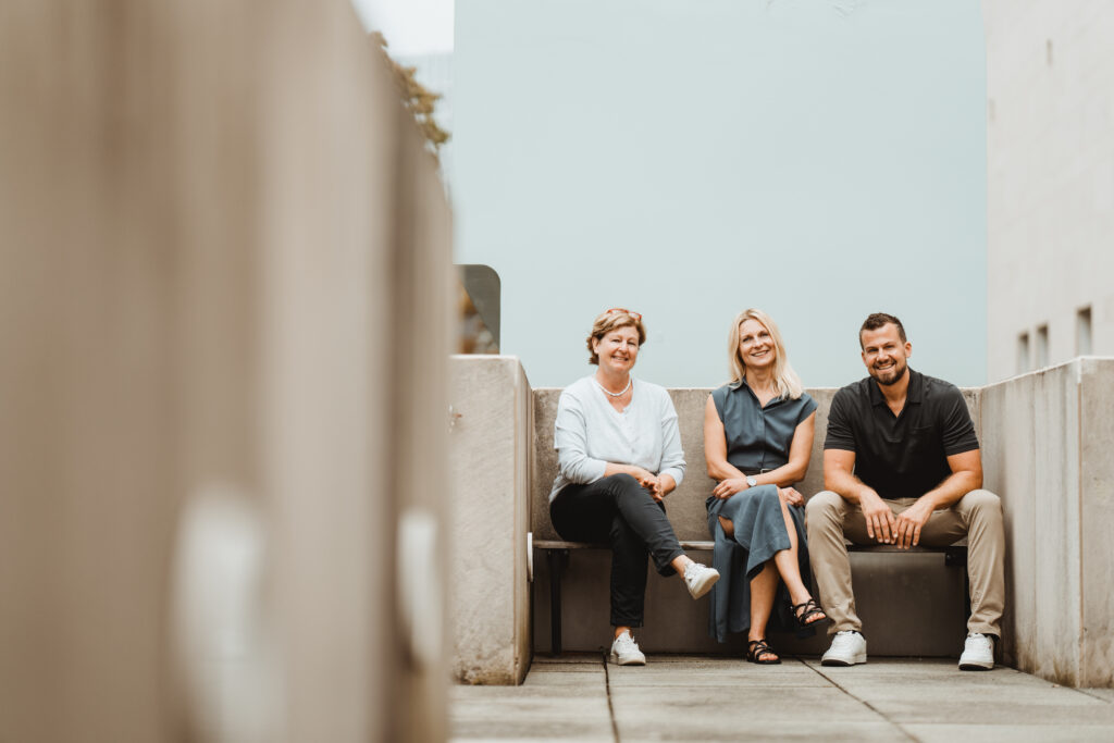 Gruppe aus zwei Frauen und einem Mann auf Sitzbank in urbaner Architektur mit hellem Hintergrund